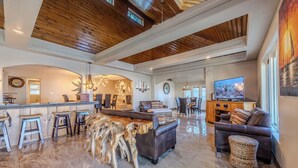 A spacious living area with a wooden ceiling, a bar area with stools, a leather sofa, and a television.