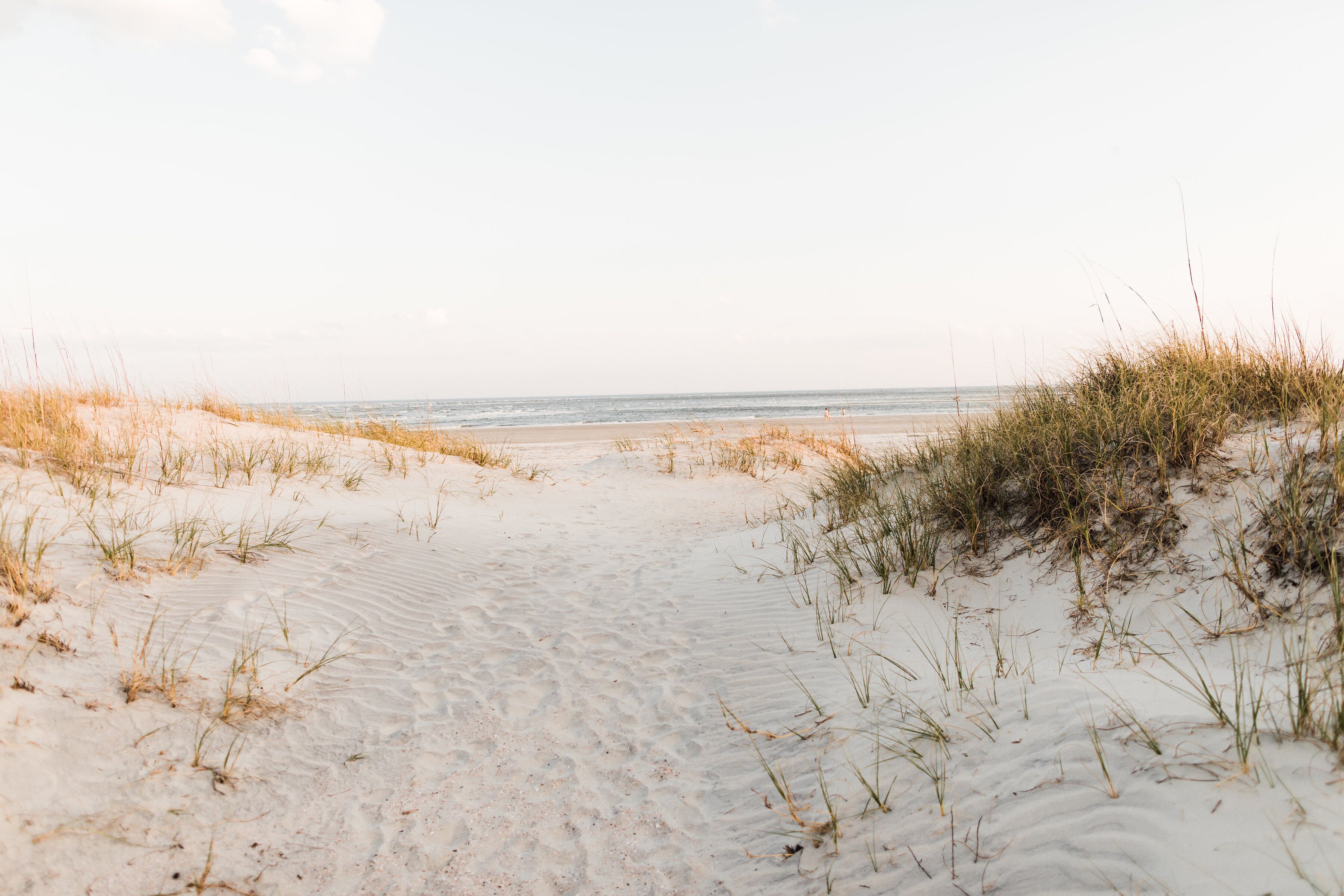 Ein sandiger Küstenpfad führt zum Meer, begleitet von Dünen an beiden Seiten.