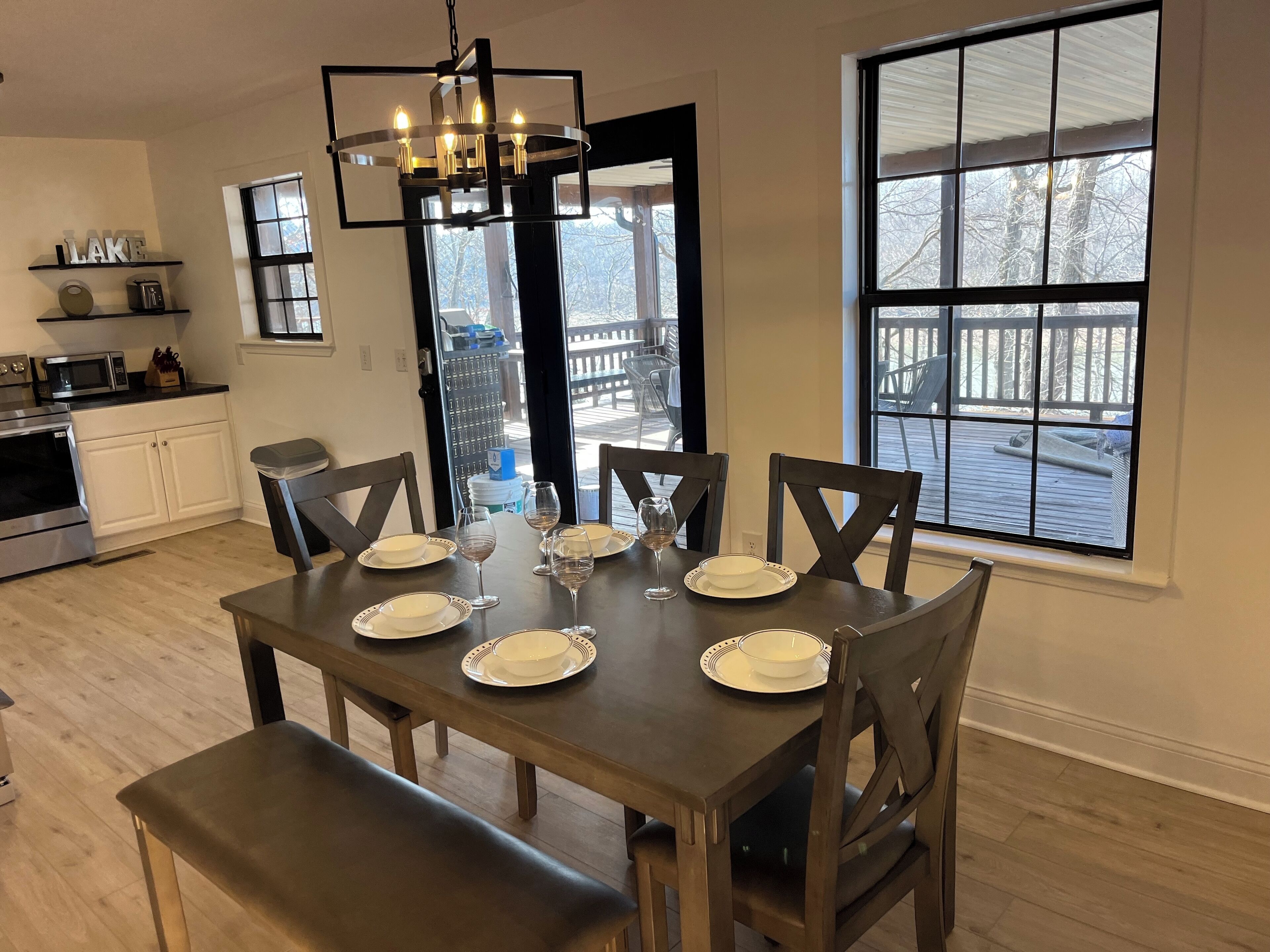 Un comedor con una mesa de madera oscura dispuesta para cuatro personas, rodeada de sillas. Un candelabro cuelga encima, y un ventanal ofrece vistas a una terraza y árboles.