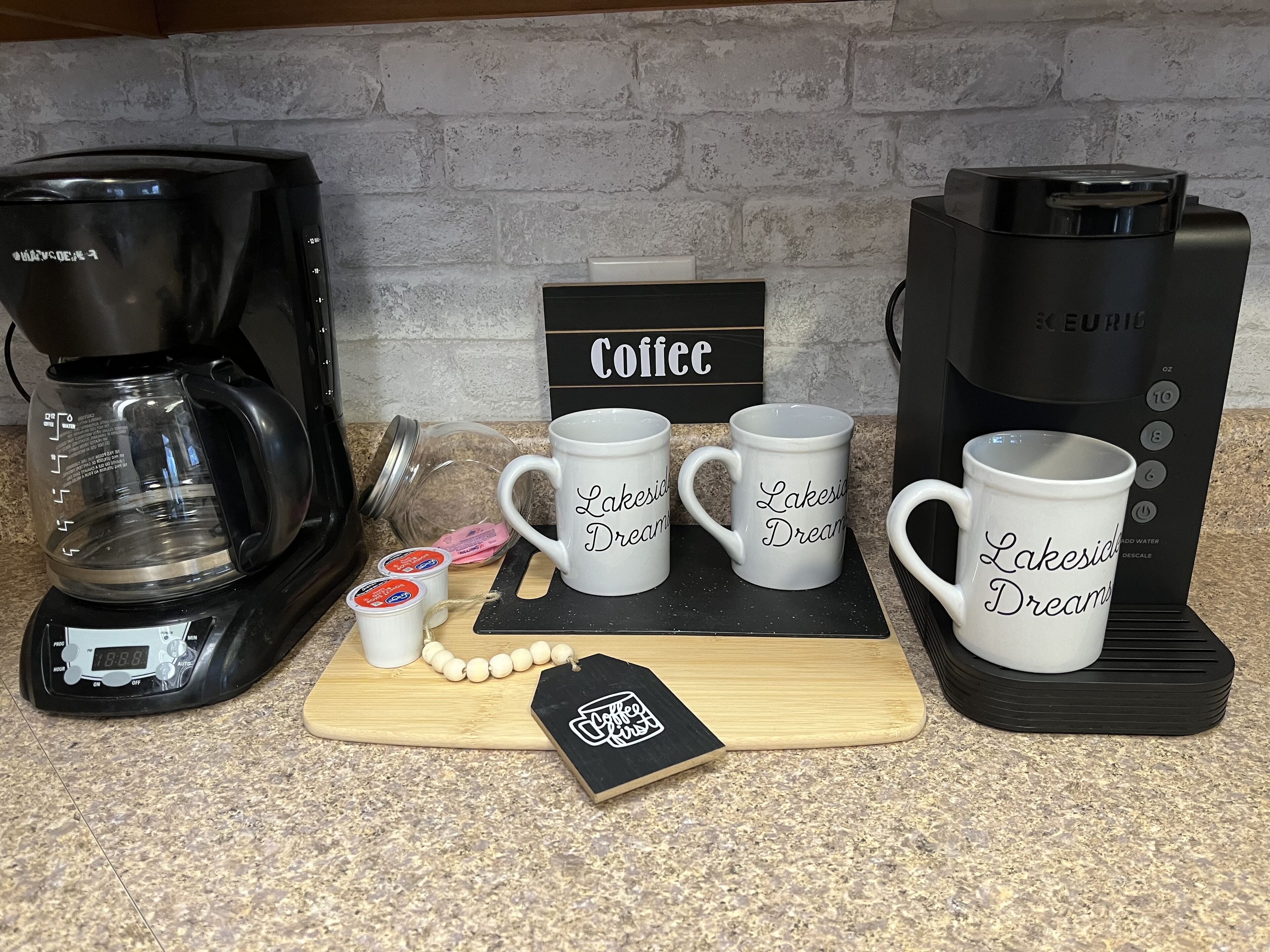 A coffee station with a coffee maker, two white mugs with 'Lakeside Dreams' text, a coffee pod, and a wooden board with a coffee mug and a tag.