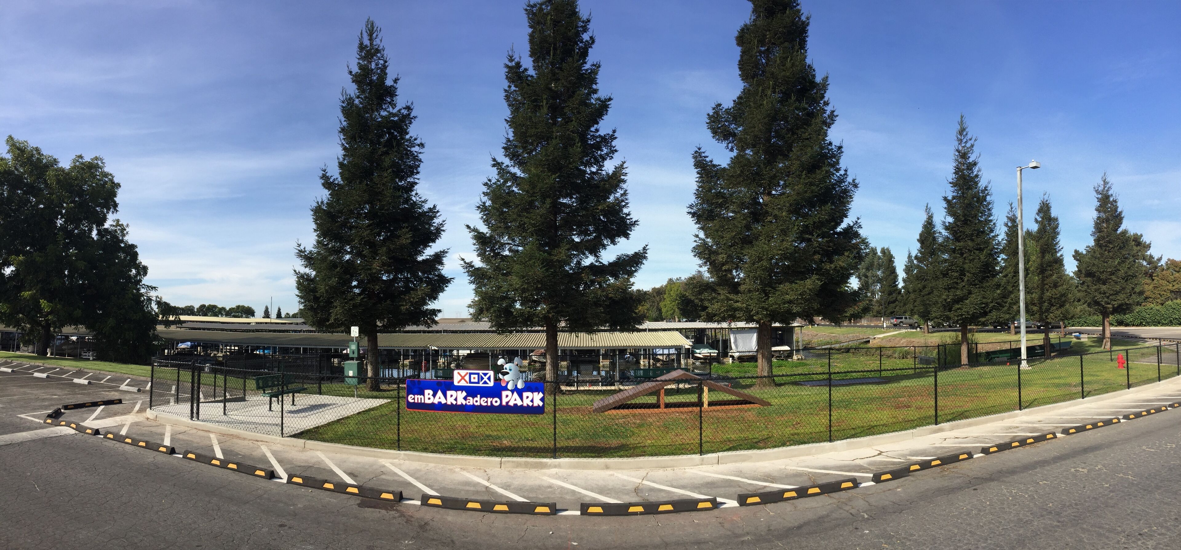 Un'area verde recintata con un cartello per emBARKadero Park, un parcheggio e alberi alti.
