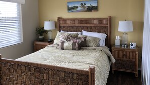 A bedroom with a bed, two bedside tables, a window with blinds, a wall-mounted painting, and a nightstand with a lamp.