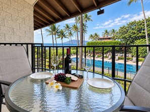 A glass-top table with a bottle of wine, a glass, and a fruit platter on a balcony overlooking a pool and palm trees.