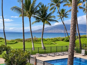 A pool area with lounge chairs, palm trees, and a view of the ocean and wind turbines.