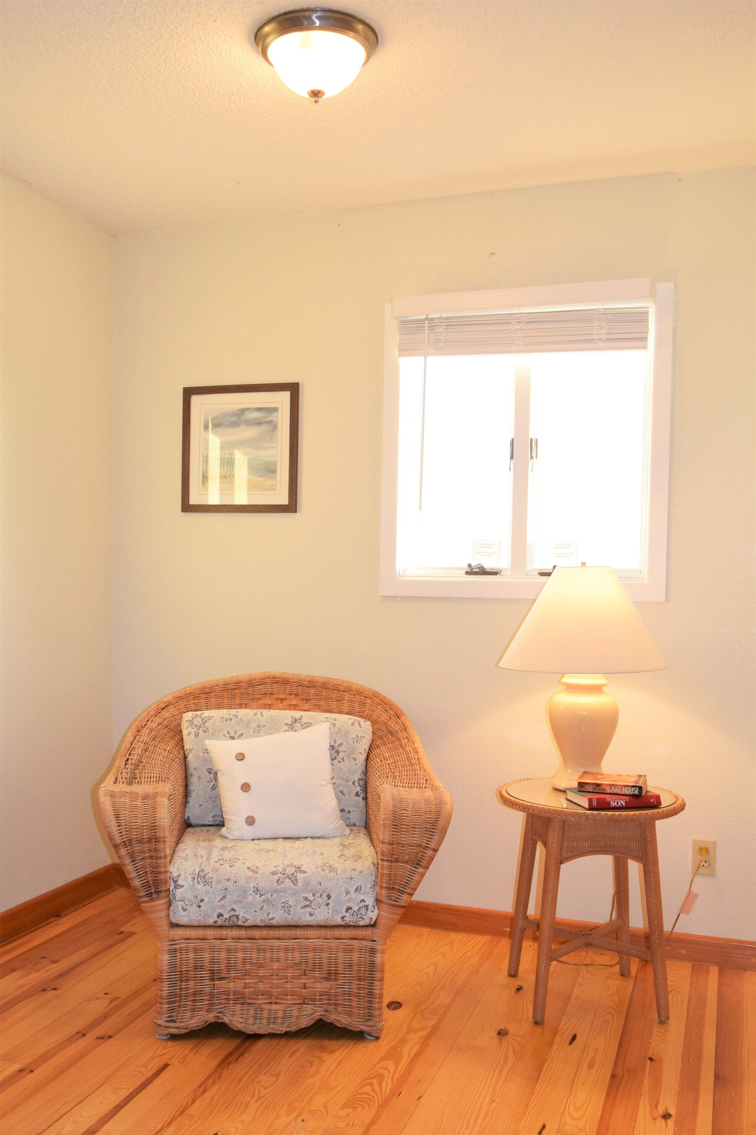 Quarto com cadeira de vime, uma pequena mesa com abajur, um quadro na parede e uma janela com persianas.