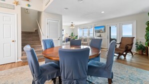 A spacious living area with a round wooden table, blue upholstered chairs, a large window with a view, and a staircase leading to another level. - "Oceans Edge" - Luxury Oceanfront home, 4 Bedrooms / 2.5 Bathrooms (Holden Beach)