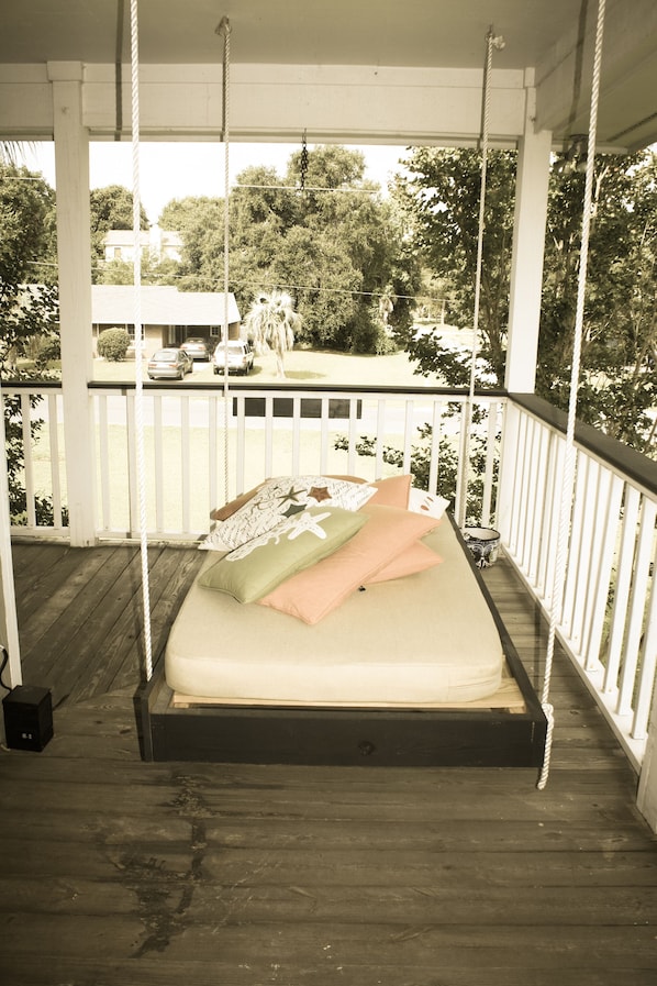 Ein Bett an einer Schaukel auf einer überdachten Veranda mit Blick auf ein Haus und Autos im Hintergrund.