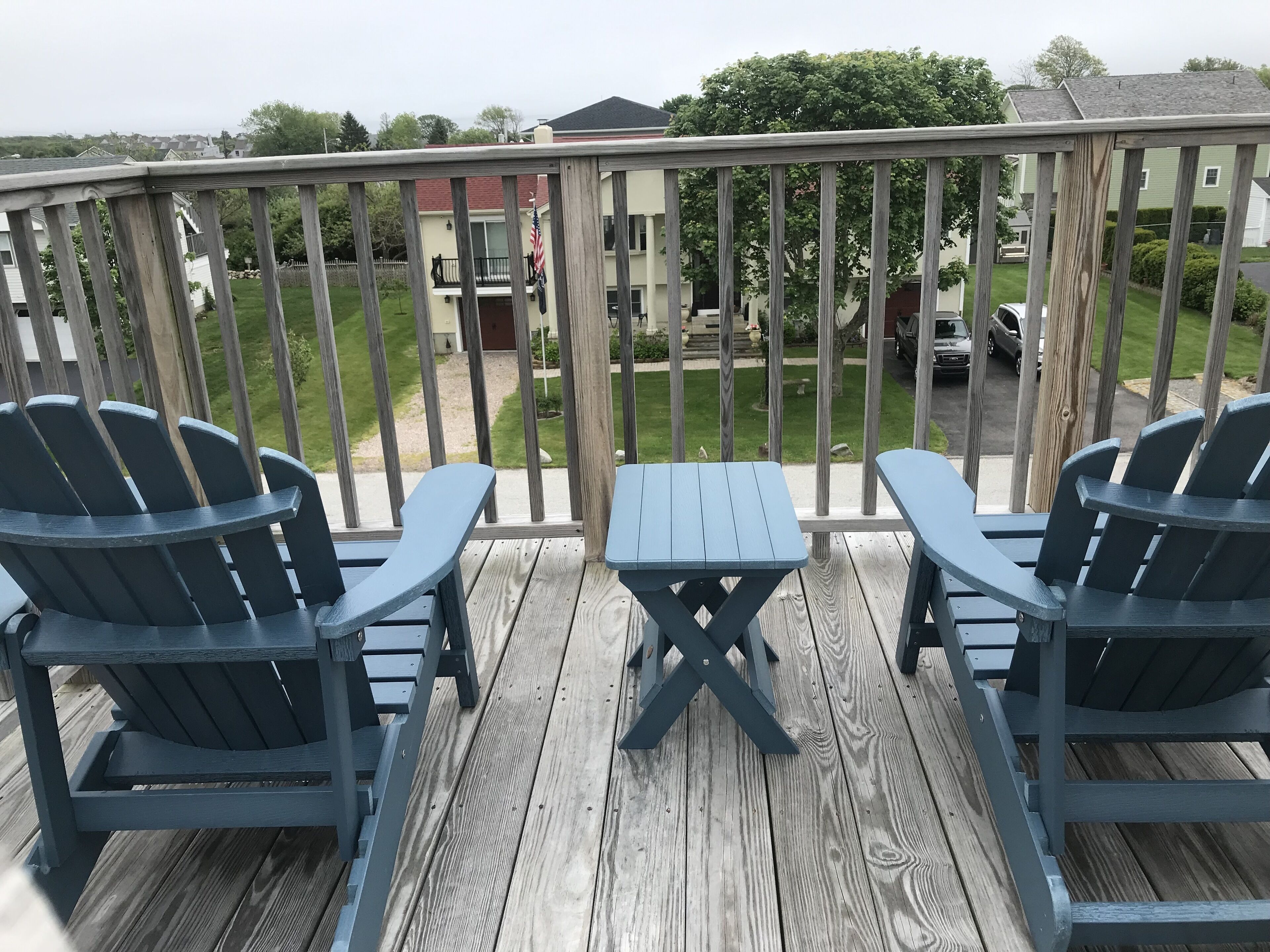 A wooden deck with two Adirondack chairs and a small table, overlooking a residential area with houses and a lawn.