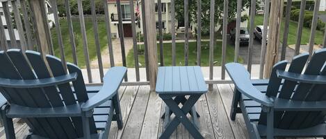 A wooden deck with two Adirondack chairs and a small table, overlooking a residential area with houses and a lawn.