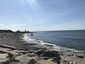 Eine felsige Küstenlinie mit brechenden Wellen, ein Leuchtturm und ein Gebäude in der Ferne.