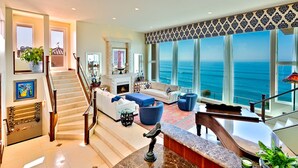 A spacious living area with a fireplace, a large window overlooking the ocean, and a staircase leading to an upper level. - Oceanfront Beach, Pool, Hot Tub, Pickleball, Movie Theater, Elevator & Golf Cart (La Jolla)