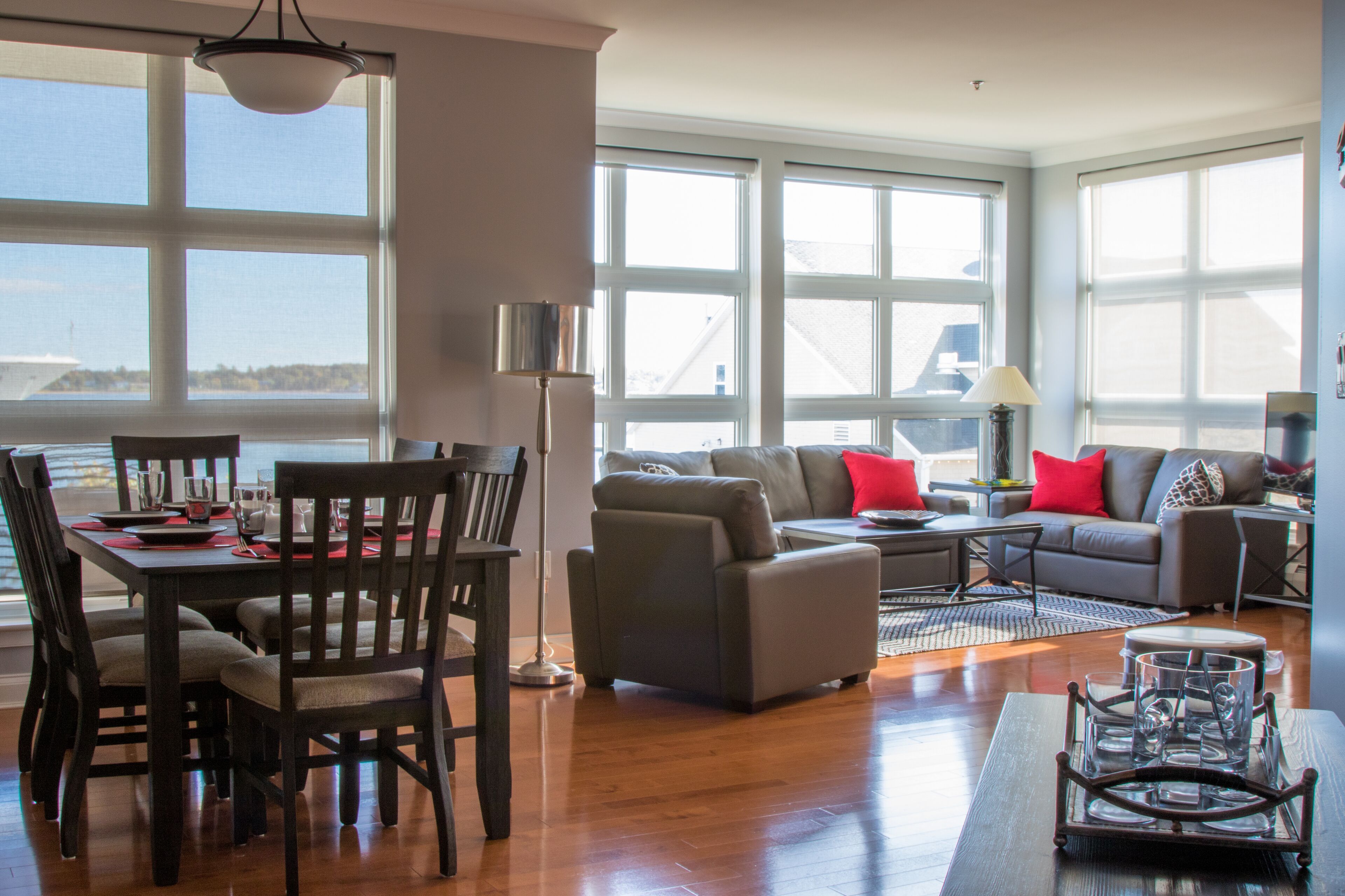 A spacious living area with a dining table, chairs, sofas, and a coffee table. Large windows provide ample natural light and a view of the outdoors.