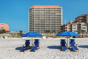 A beachfront with blue beach chairs and umbrellas, a tall hotel building, and a sandy beach. - Gulf-Front Tides Unit 202 | 2BR | Sleeps 6 (Miramar Beach)