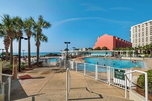 A pool area with a view of the ocean, surrounded by palm trees and a building with balconies. - Gulf-Front Tides Unit 202 | 2BR | Sleeps 6 (Miramar Beach)