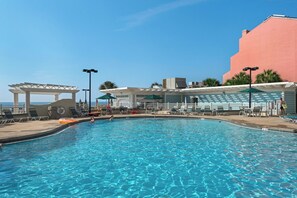 A swimming pool with a clear blue water surface, surrounded by lounge chairs and umbrellas, and a building with a pink facade in the background. - Gulf-Front Tides Unit 202 | 2BR | Sleeps 6 (Miramar Beach)