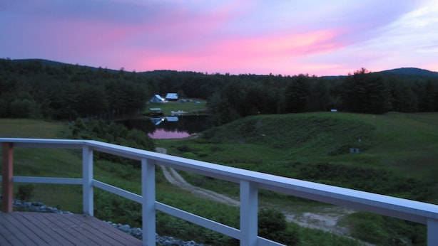 A view from a balcony overlooking a lake with a house in the distance, surrounded by forested hills. - Beautiful Farmhouse located on 1200 acres of private bliss with a quiet pond. (Brownfield)