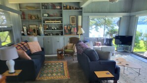 A living room with a sofa, armchairs, a coffee table, and a TV. There is a bookshelf with various items and a window with a view of greenery. - Quiet, Private Lakefront Cottage - Skaneateles Lake (Homer)