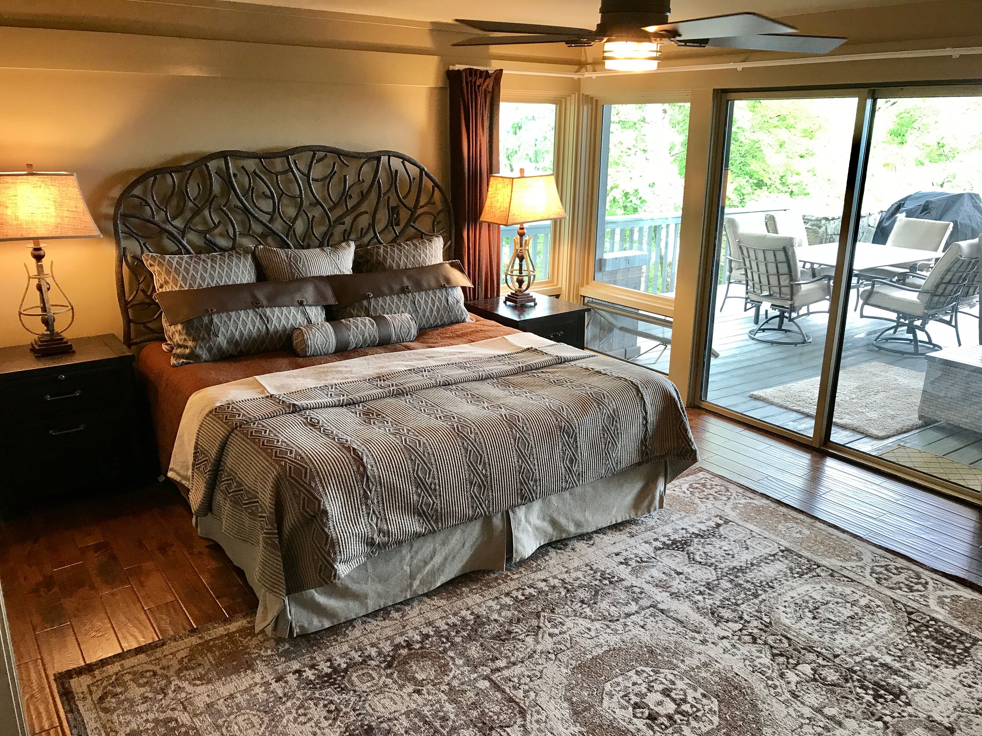 A bedroom with a large bed, a nightstand, a lamp, and a view of an outdoor patio with chairs and a table.