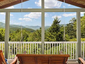 A porch with a swing chair and a bench, overlooking a forested mountain range.