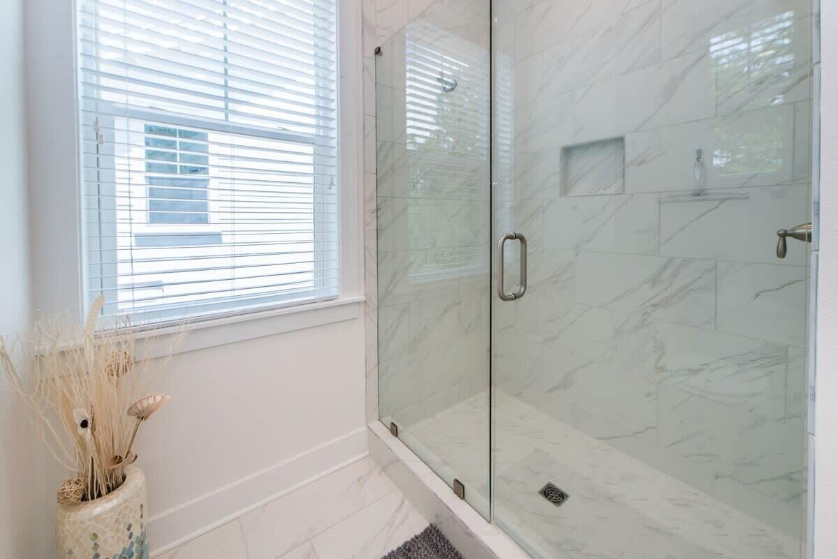 A glass-enclosed shower with a marble wall, a window with blinds, and a vase with dried plants.