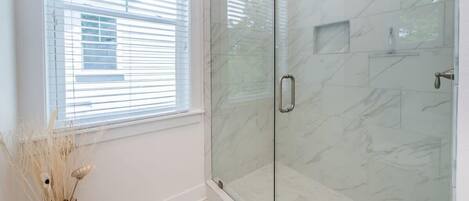 A glass-enclosed shower with a marble wall, a window with blinds, and a vase with dried plants.