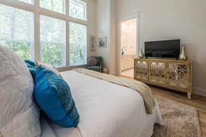 Une chambre avec un grand lit, un téléviseur posé sur un meuble en bois et une fenêtre équipée de stores.