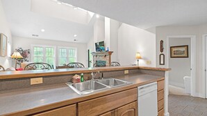 A kitchen with a large island, stainless steel sink, and a dishwasher. There are two windows with white frames, a door, and a mirror above the sink.