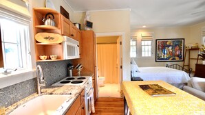 A compact kitchen with a sink, stove, and wooden cabinets. A bed with a yellow bedspread is visible in the adjacent room. - In the Heart of Rosemary Beach - Beach, Pool, & Shops Steps Away (Rosemary Beach)