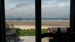 A beach view from inside a house with a patio table and chairs, a wooden deck, and a view of the ocean and a distant island. - Oceanfront, Amazing View, Hot Tub Facing Ocean (Arch Cape)