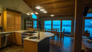 A modern kitchen with wooden cabinets, a central island, and a view of the ocean through large windows. - Oceanfront, Amazing View, Hot Tub Facing Ocean (Arch Cape)