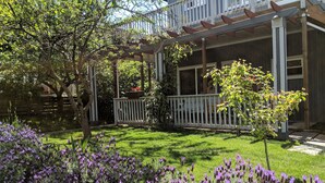 Una casa con porche cubierto, patio cercado y flores moradas.
