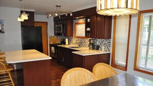 Una cocina moderna con armarios de madera oscura, encimera blanca y estufa integrada. Hay una zona de comedor con sillas de mimbre y una mesa.