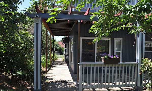 A covered porch with a white railing, a pathway leading to a house, and a garden area with plants and a wooden structure.