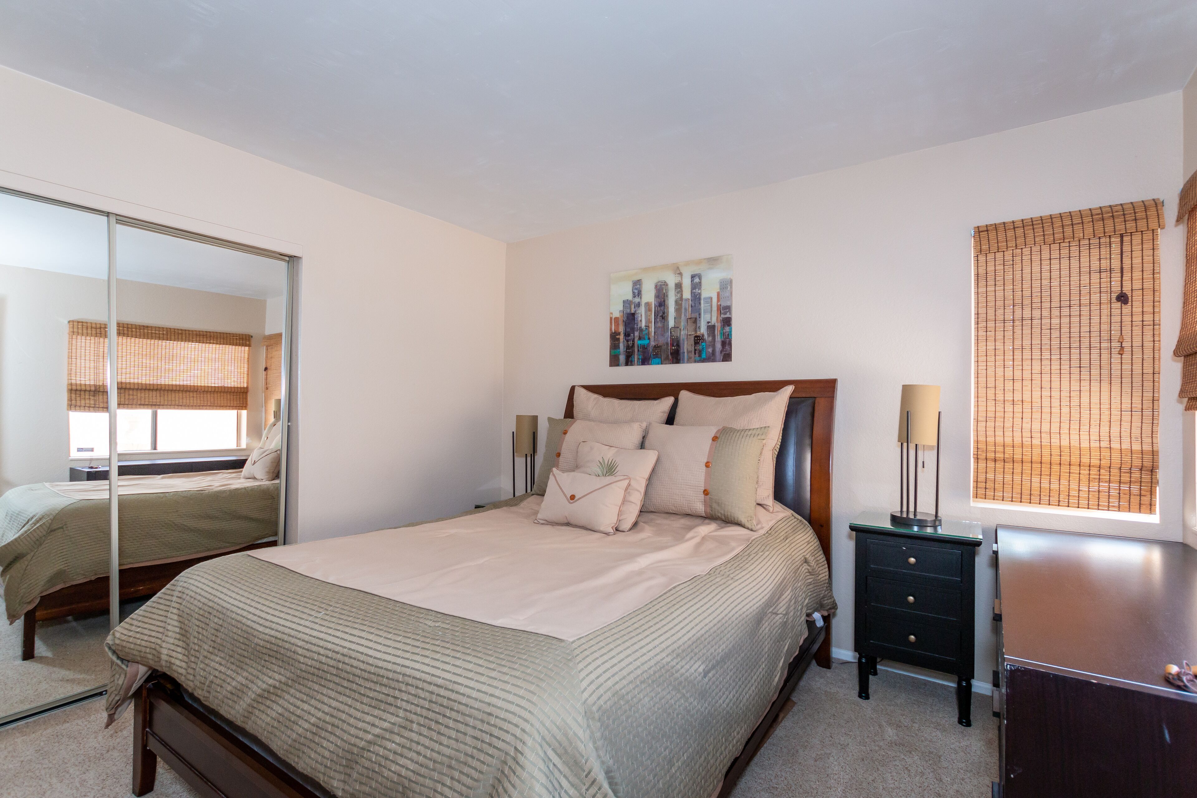 A bedroom with a bed, a bedside table, a mirror, a window with blinds, and a wall-mounted artwork.