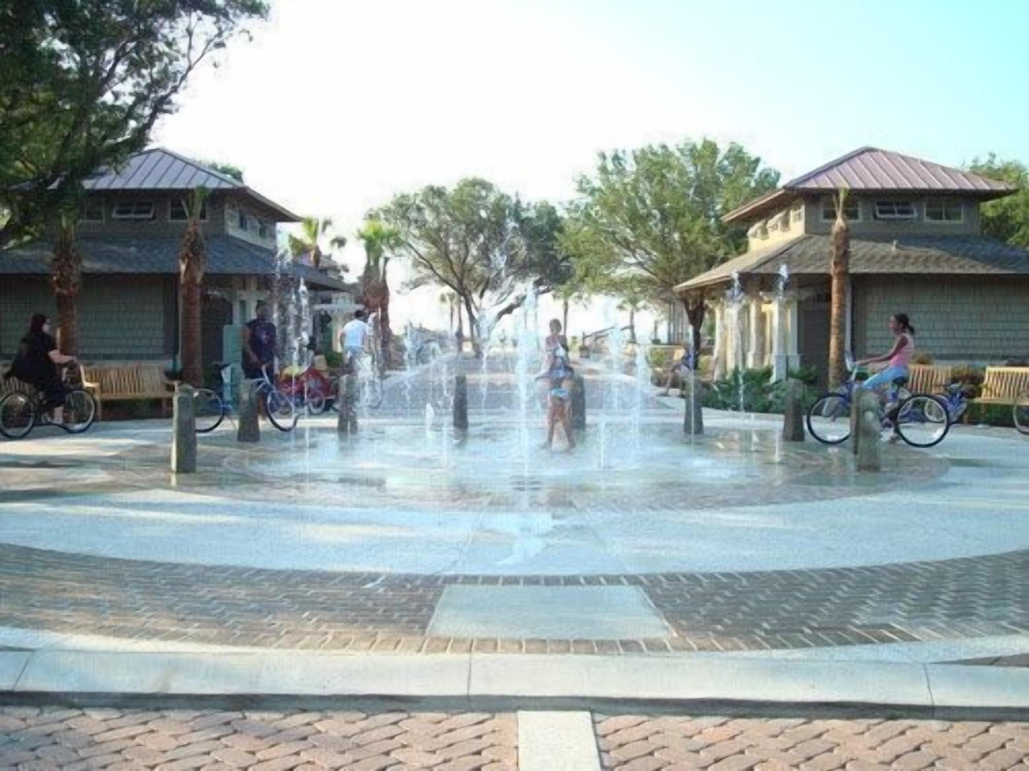 Ein öffentlicher Platz mit einem zentralen Wasserspiel, umgeben von Gebäuden mit Ziegeldächern und Bänken.