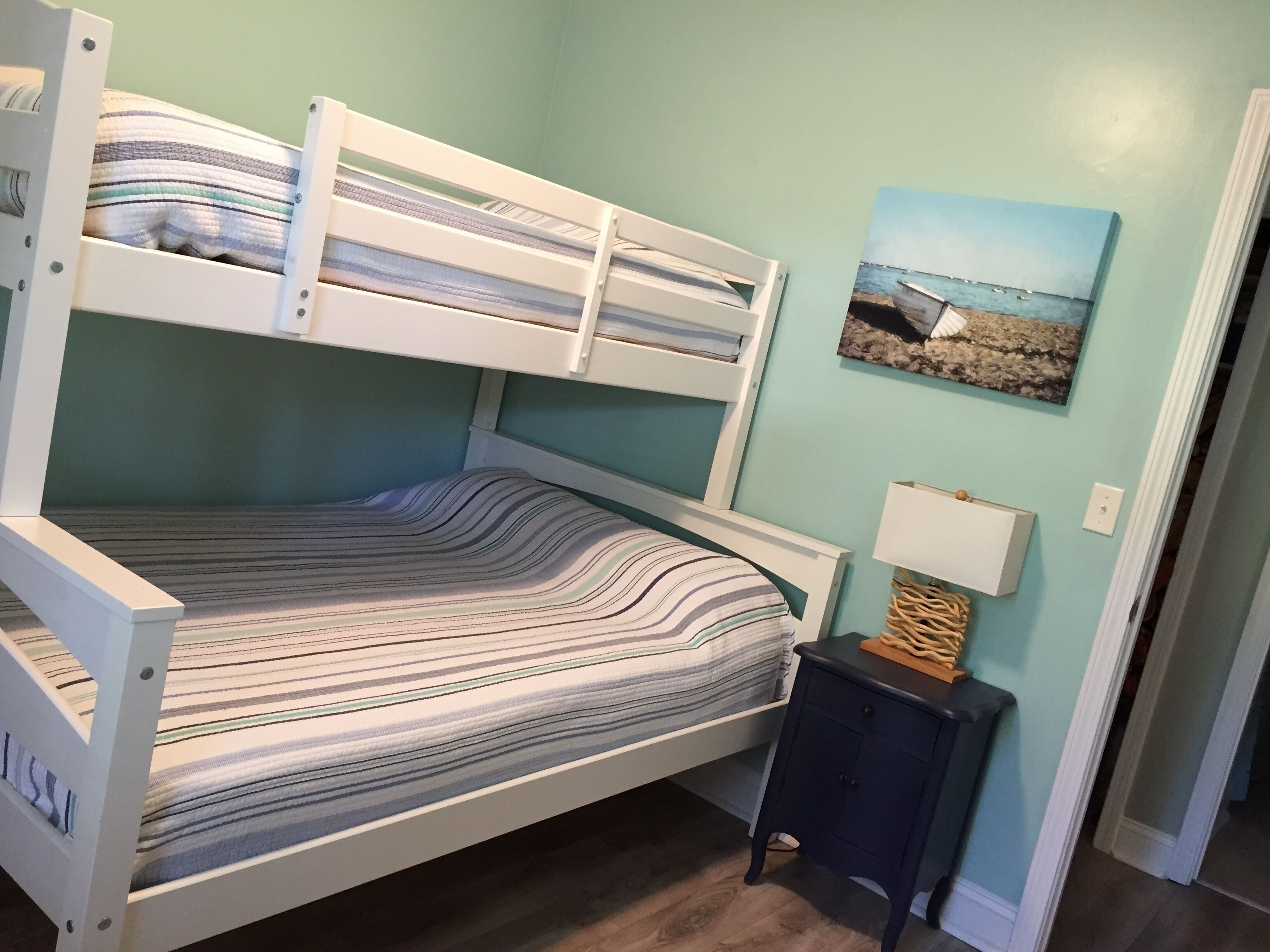 Een stapelbed met gestreepte lakens, een klein nachtkastje met een lamp, en een schilderij van een strand.