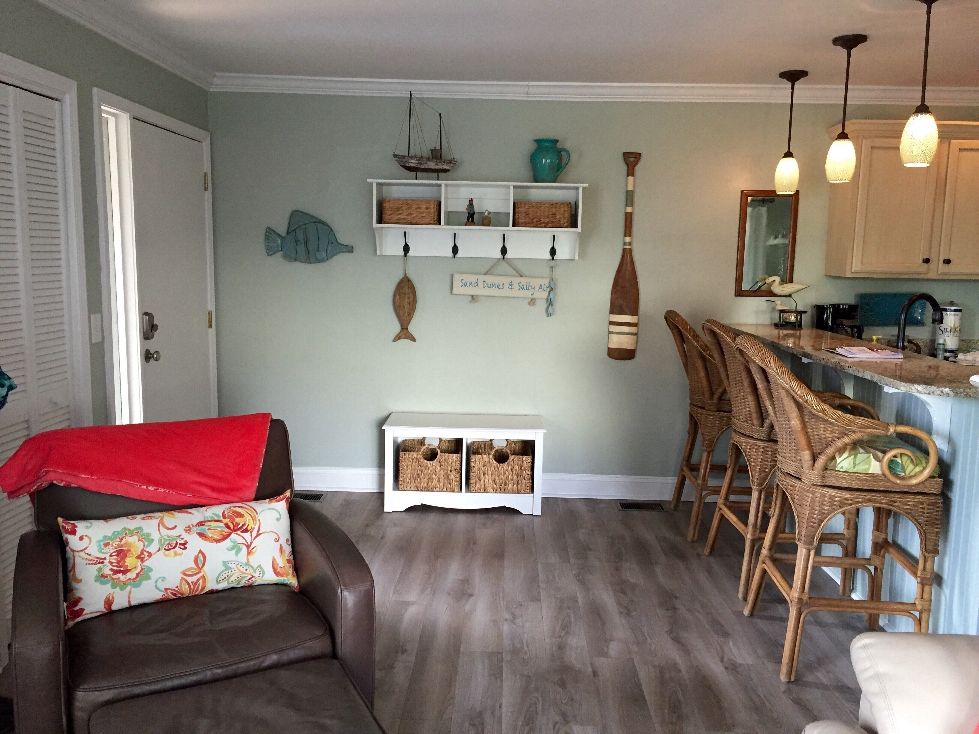 A living room with a brown leather sofa, a wicker chair, a small white console table, and a kitchen area with wooden cabinets and a countertop.