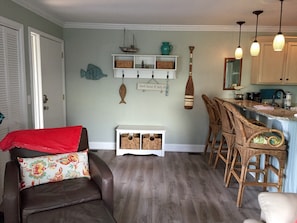 Un salon avec un canapé en cuir marron, un fauteuil en osier, une petite table console blanche et un coin cuisine doté de meubles en bois et d’un plan de travail.