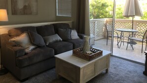 Un salón con un sofá gris oscuro, una mesa de centro blanca y vistas a un patio exterior con una mesa y sillas.