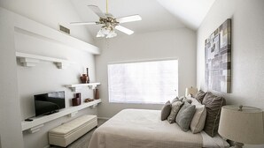 Un dormitorio con cama, televisión, ventilador de techo, ventana y cuadros en la pared.