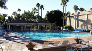 Un área de piscina con sillones, una zona de estar cubierta y palmeras al fondo.