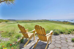 Twee houten Adirondack stoelen op een verhard gebied, met een grasveld en een uitzicht op de oceaan op de achtergrond.