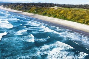 Vista costera con olas rompiendo sobre una playa arenosa, un acantilado boscoso y un edificio en la distancia.