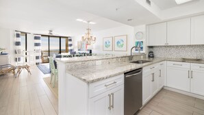 A modern kitchen with white cabinets, a granite countertop, and a dishwasher. There is a dining area with a table and chairs, and a view of the city through large windows.