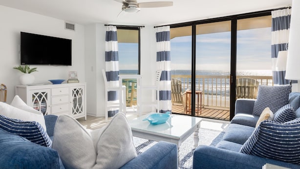 A modern living room with a large flat-screen TV, a ceiling fan, and a view of the ocean through large sliding glass doors.