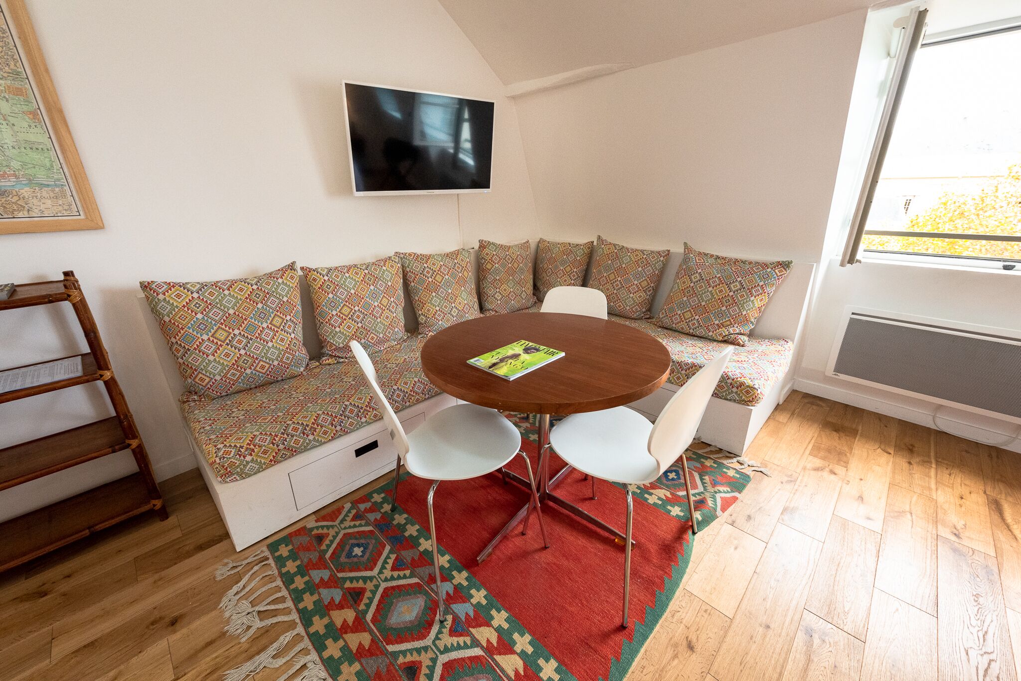 Un salon chaleureux avec une table ronde en bois, des chaises blanches, un canapé à motifs et un téléviseur fixé au mur.