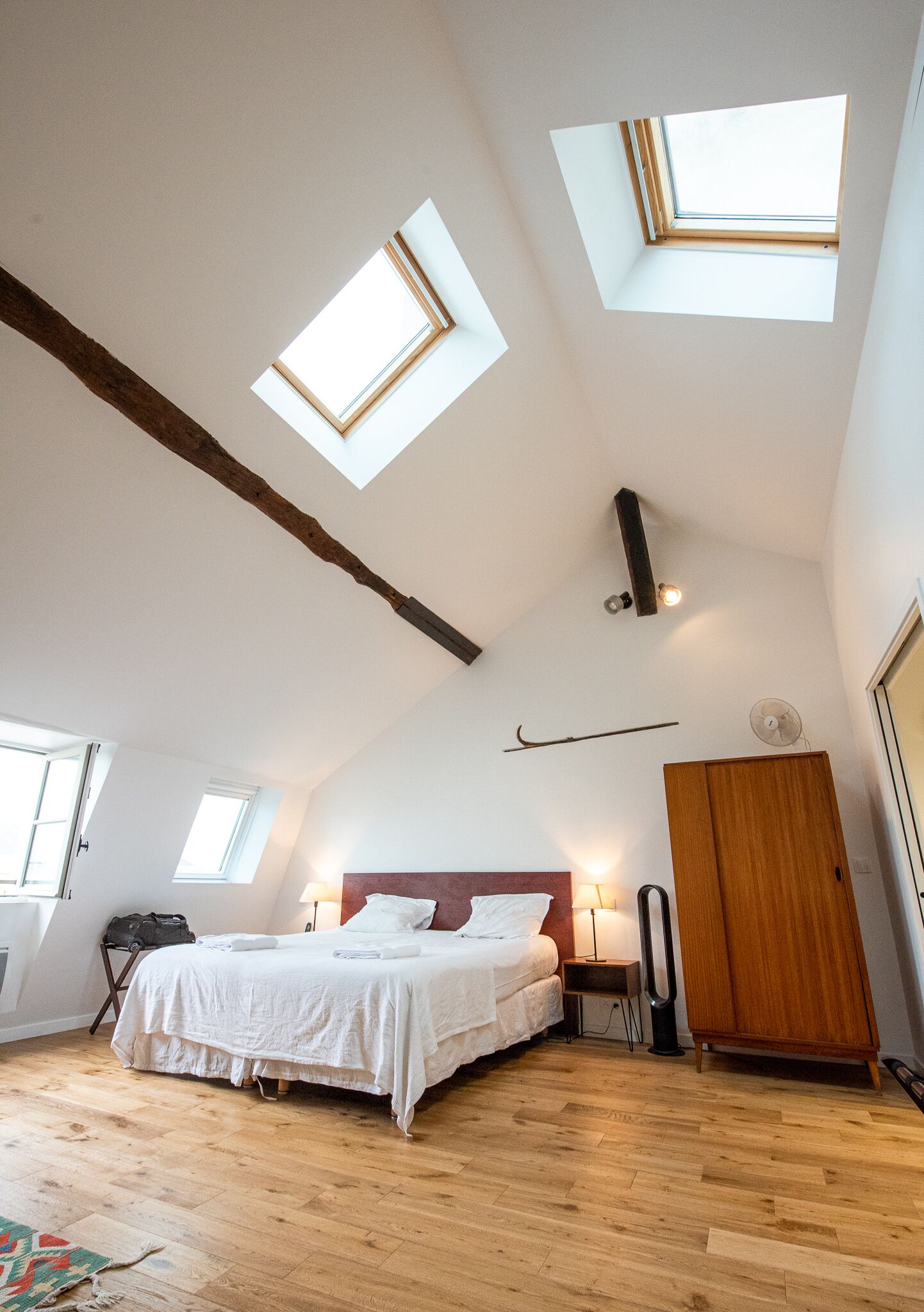 Ein Schlafzimmer mit schrägem Dach, Dachfenstern, einem Bett, Nachttischlampen, einem Kleiderschrank und einem kleinen Tisch.