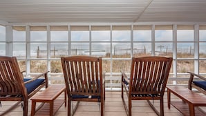 Una terraza techada con tres sillas de madera y una mesa, con vista a una playa con valla de madera y dunas cubiertas de pasto.