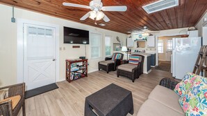 Una sala de estar acogedora con ventilador de techo, televisor, mobiliario de mimbre y una zona de cocina con refrigerador.
