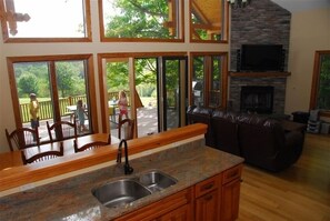Relax in this spacious living area with a kitchen counter, stone fireplace, and large windows offering a view of the deck and surrounding greenery.
